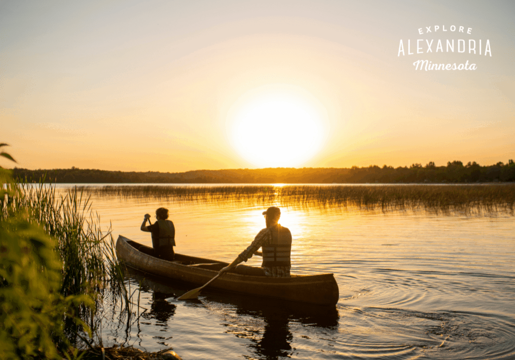 Two people paddle a canoe on a calm lake at sunset near Alexandria, surrounded by reeds and trees, with text in the sky that reads, "Explore Alexandria Minnesota—perfect for nature lovers and trap shooters alike.
