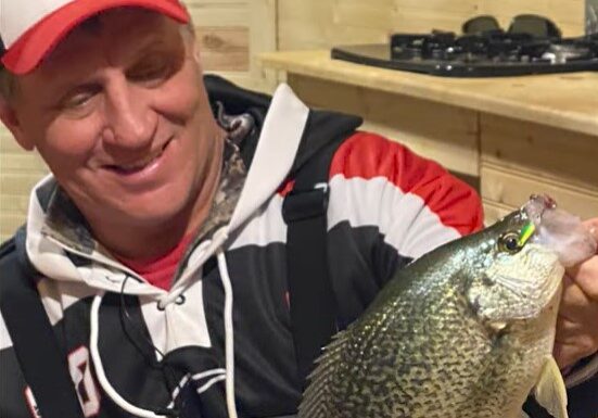 A smiling man in a red and white cap holds up a large crappie fish indoors, wearing a black and red jacket. Light wooden walls and a stove are visible in the background—a perfect moment to share your favorite fishing tip.
