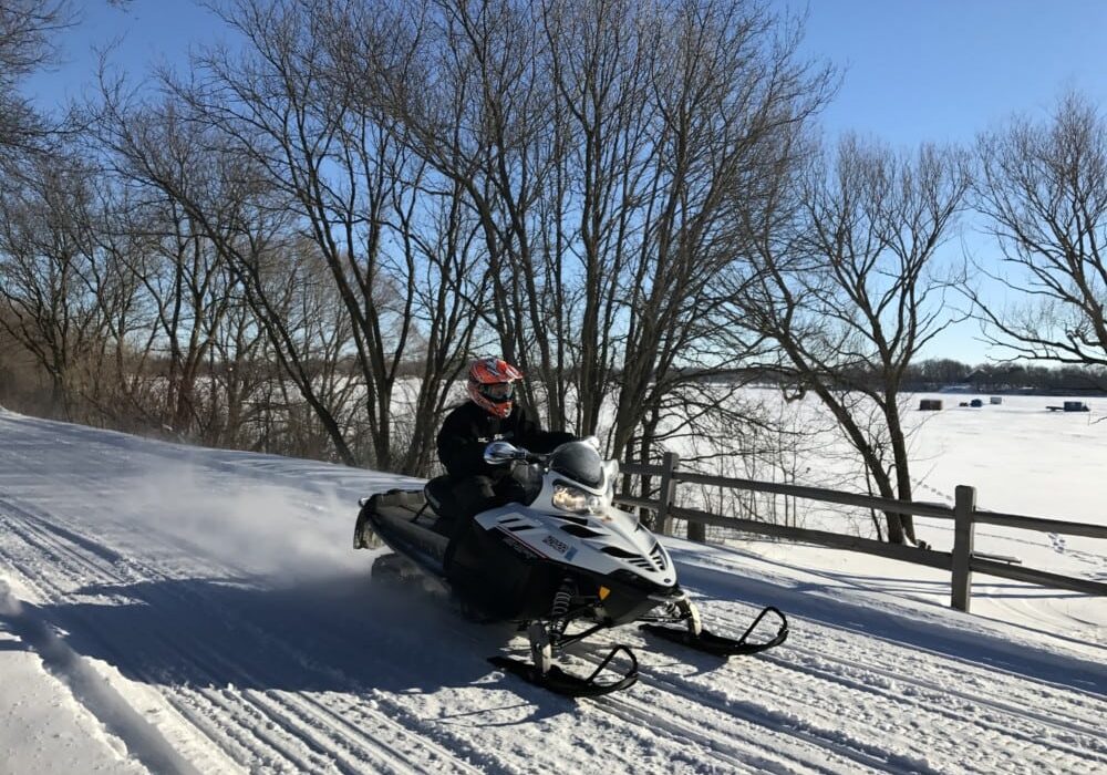 A person in winter gear rides a snowmobile along Alexandria snowmobile trails, gliding past a wooden fence with bare trees and a snowy landscape in the Alexandria area under a clear blue sky.