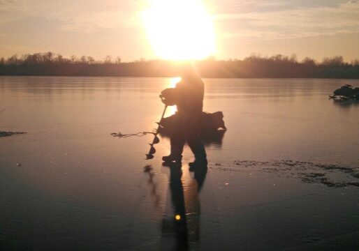 A person stands on a frozen lake at sunset, holding ice fishing equipment. Their silhouette is reflected on the ice, with another figure and equipment in the background—capturing the excitement of a first ice fishing report.