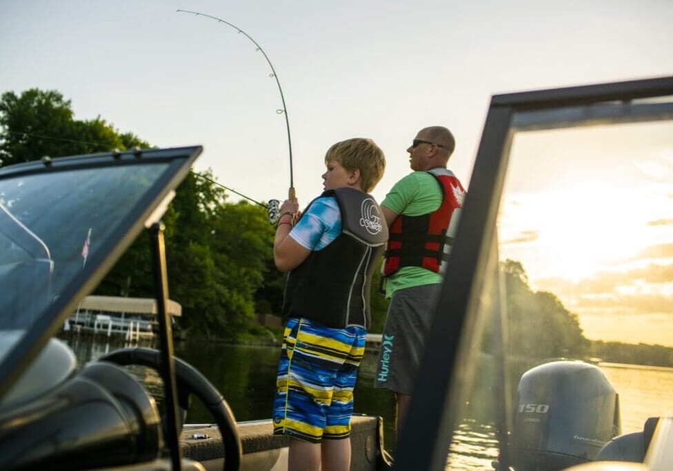 A boy and an adult wearing life jackets fish from a boat on a calm lake at sunset during their Alexandria getaway, with trees and a dock visible in the background.