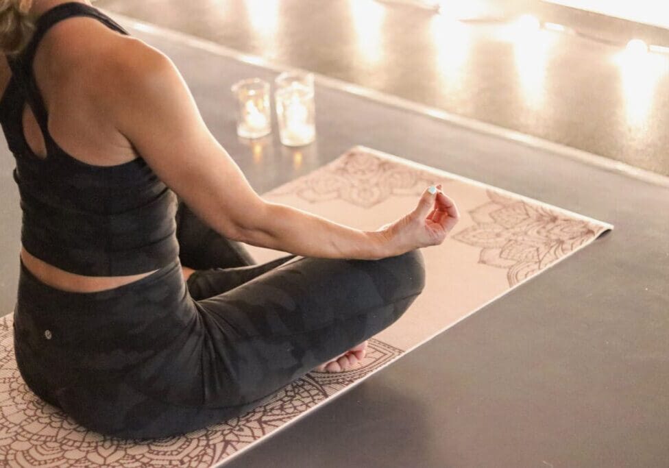 A person in black athletic wear sits cross-legged on a patterned yoga mat, meditating with one hand resting on their knee at Focus Fitness Yoga Studio. Soft candlelight glows in the background, creating a peaceful atmosphere.