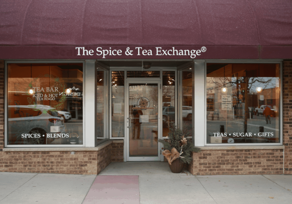 The Spice & Tea Exchange storefront features a maroon awning, glass door, and large windows showcasing signs for tea, spices, blends, sugar, and gifts. A decorative plant greets visitors at the entrance.