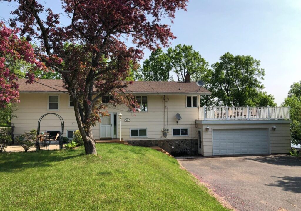 A beige split-level house with a two-car garage, white balcony railing, and a satellite dish. A large tree with red leaves stands out front—green grass surrounds the drive. Memories are Made on Brophy Shores in this inviting home.