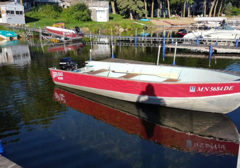 A small red and white motorboat labeled "POLICE RESCUE" is docked in a marina on calm water at Geneva Beach Resort, with rentals, other boats, docks, and lakeside buildings visible. A person’s shadow is cast on the boat.