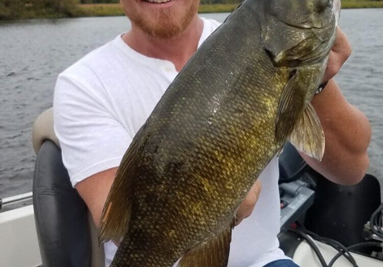 A smiling man wearing sunglasses and a cap sits in a boat on Alexandria Lakes, proudly holding up a large Smallmouth Bass he caught. Trees and cloudy skies are visible in the background.