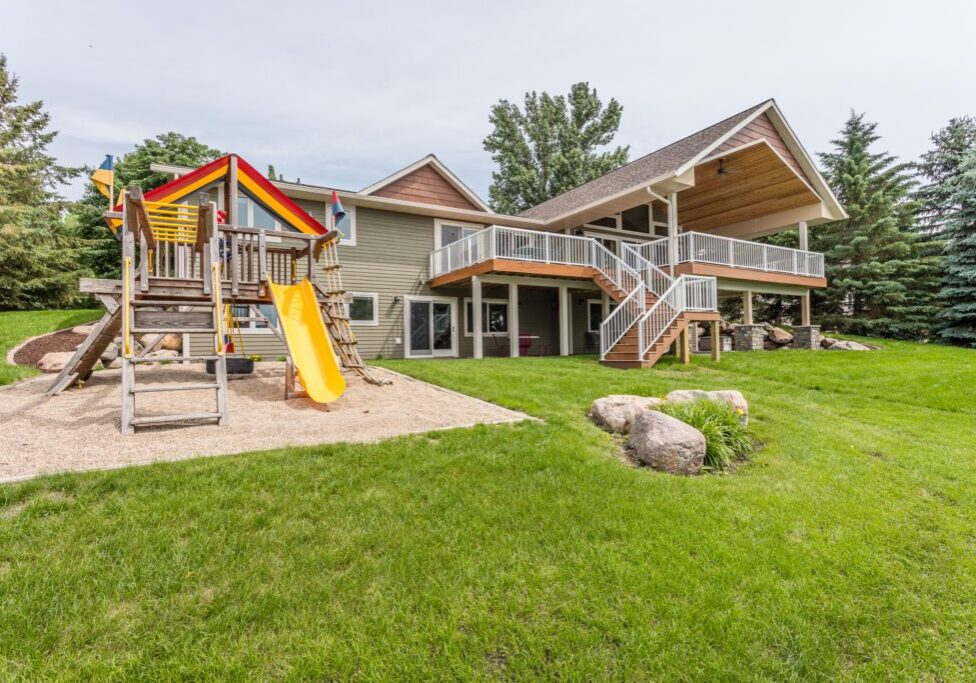 The Haven is a modern two-story house with a large covered deck, white railings, and an outdoor staircase. In the yard, a wooden playset with swings and a yellow slide sits on gravel, surrounded by green grass and trees.