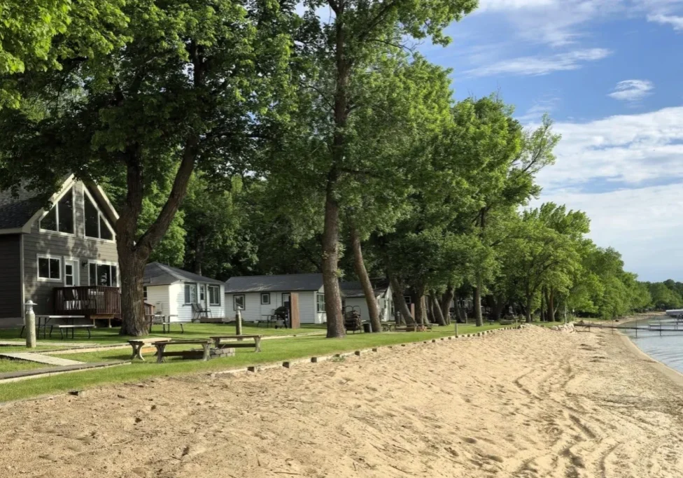 Several cottages and cabins, shaded by large trees, line the sandy lakefront at Viking Bay Resort under a partly cloudy sky, with docks and calm water visible on the right side.