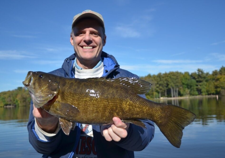 A smiling person wearing a cap and jacket holds up a large bass, celebrating Bass Season in front of a calm lake with trees and a blue sky in the background.