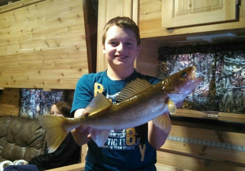 A smiling boy in a blue shirt proudly holds his catch for big fish pictures inside a wood-paneled room, possibly a cabin, while another person relaxes on a couch in the background.