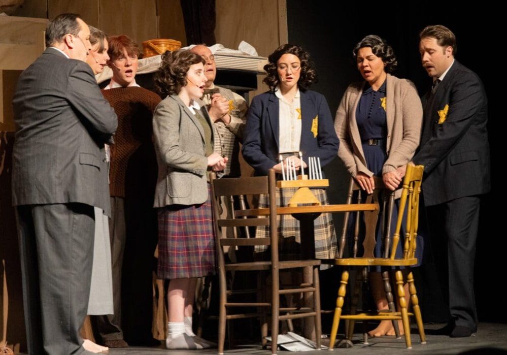 A group of actors in period clothing stands closely together around a table on the Andria Stage, appearing serious and contemplative—some wear yellow Star of David patches, evoking a powerful scene from The Diary of Anne Frank.