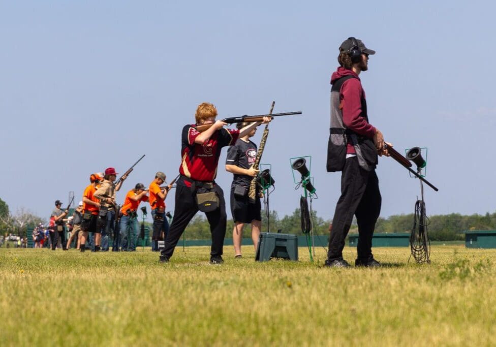A group of people stands in a row outdoors near Alexandria, aiming shotguns at targets. Some wear ear and eye protection, and the setting is a grassy field under a clear sky.