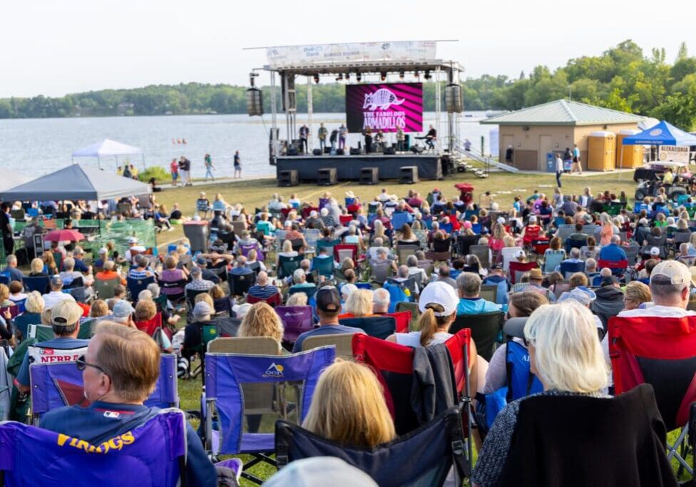 A large crowd sits in lawn chairs facing an outdoor stage by a lake, watching a live Summer Sounds performance. Trees and a small building are in the background, with a large screen on the stage under sunny, clear weather.