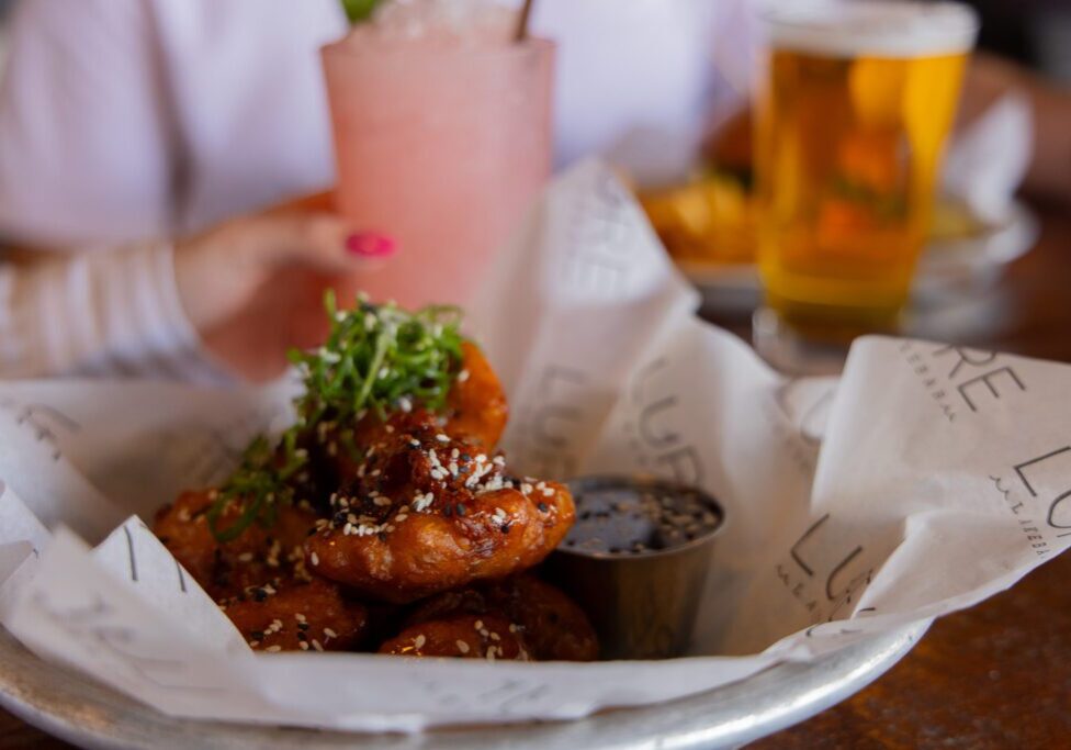 A plate of glazed fried chicken garnished with greens and sesame seeds, served with dipping sauce; a person holds a pink drink in the background at a popular Alexandria MN restaurant, with a glass of beer on the table.
