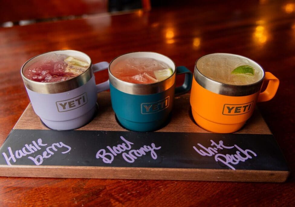 Three colorful YETI mugs on a wooden tray, each filled with a different fruity drink and labeled in chalk. This inviting setup is perfect for sharing at one of the cozy MN restaurants or while exploring Alexandria MN restaurants.