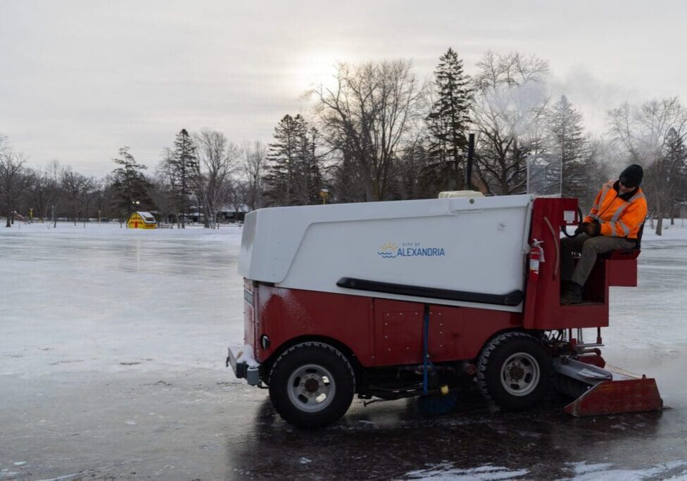 A person in an orange safety jacket drives an ice resurfacing machine across an outdoor skating rink, with bare trees, snow, and a yellow building in the background under a cloudy sky.