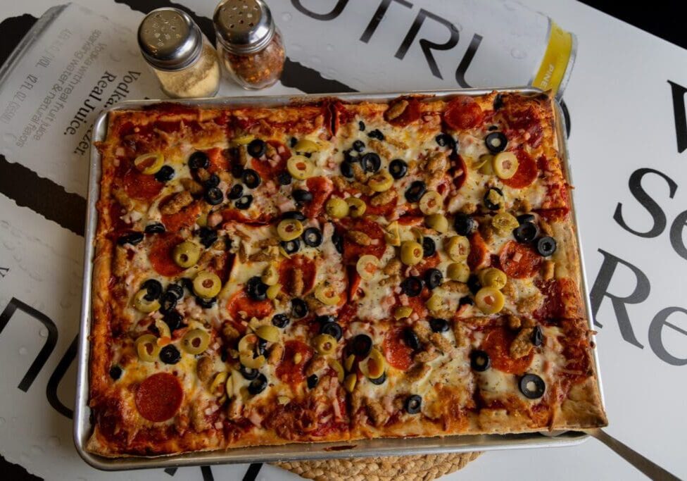 A rectangular pizza topped with pepperoni, black and green olives, and cheese sits on a tray next to shakers of grated cheese and red pepper flakes—an inviting slice of Alexandria flavor found at top Alexandria MN restaurants.