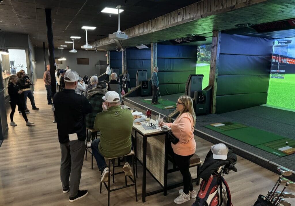 People gather at an indoor golf facility with Hook and Slice simulators, some playing and others seated at tables socializing. Golf bags are visible, and a large screen displays a golf course scene. The atmosphere is casual and lively.