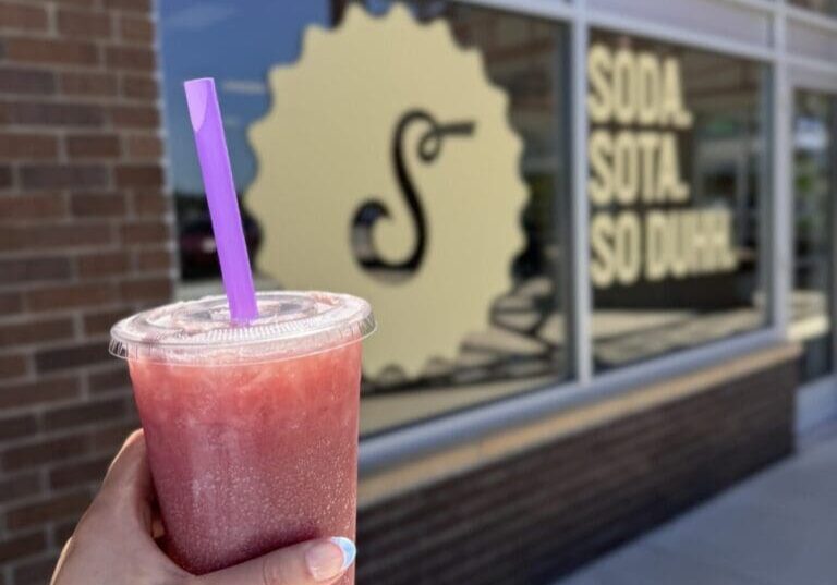 A hand with colorful nails holds a red slushy drink with a purple straw in front of Schmidty's Soda Shop window, displaying the text “SODA SOTA SO DUH!” and a stylized logo.