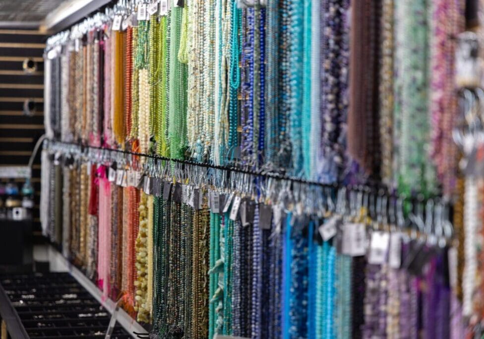 Strings of colorful beads in greens, blues, purples, pinks, and browns hang neatly organized on display racks at iTry Studios, creating a vibrant and inviting craft or bead store atmosphere.