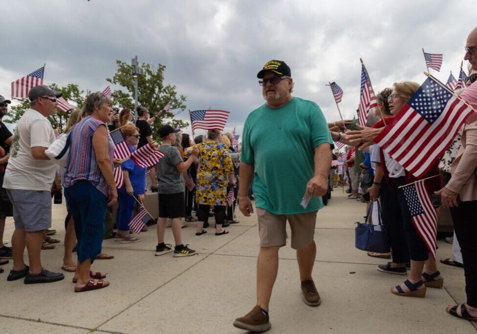 A man in a green shirt and veteran hat walks past a crowd holding American flags outdoors on a cloudy day in Alexandria. People line the sidewalk, reaching out and watching as the Vietnam Veteran receives a heartfelt Welcome Home.