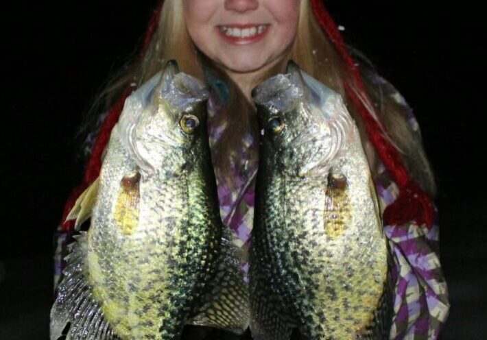 A smiling girl in a red knit hat and checkered jacket holds up two large crappie fish in both hands, standing outside at night—perfect for sharing Early January Ice Fishing success.