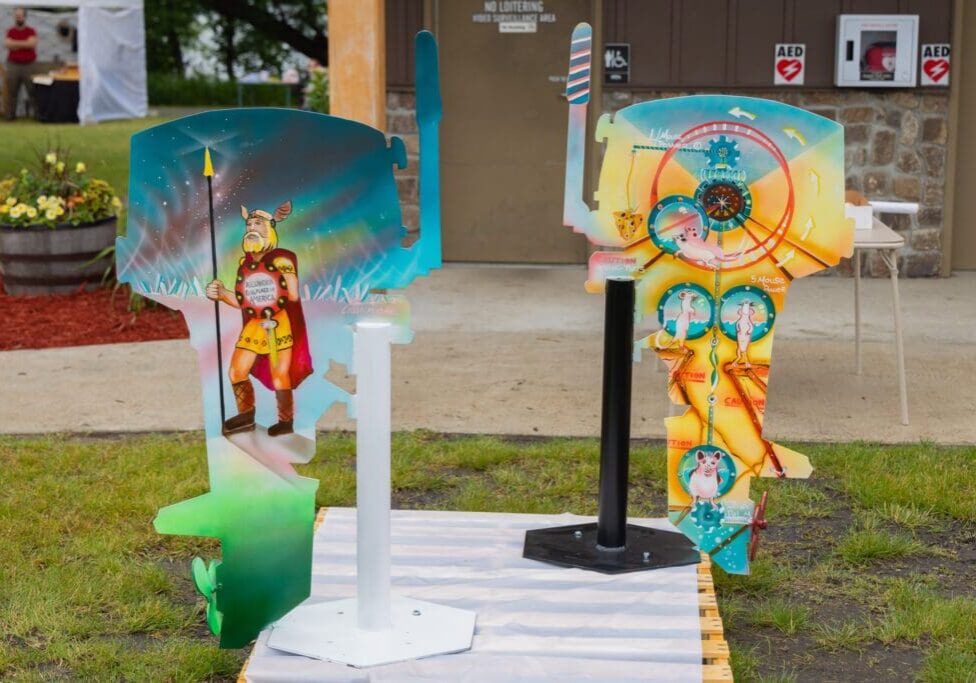 Three colorful wooden sculptures with Native American-inspired artwork stand outdoors on a mat near a building. The left sculpture depicts a warrior with a spear, while the other two feature abstract designs and faces.