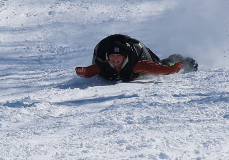 A person wearing a winter hat and gloves slides down a snowy hill face-first on a black sled, smiling as snow kicks up behind them. Bright sunlight highlights the wintry scene—perfect for skiing in Alexandria MN.
