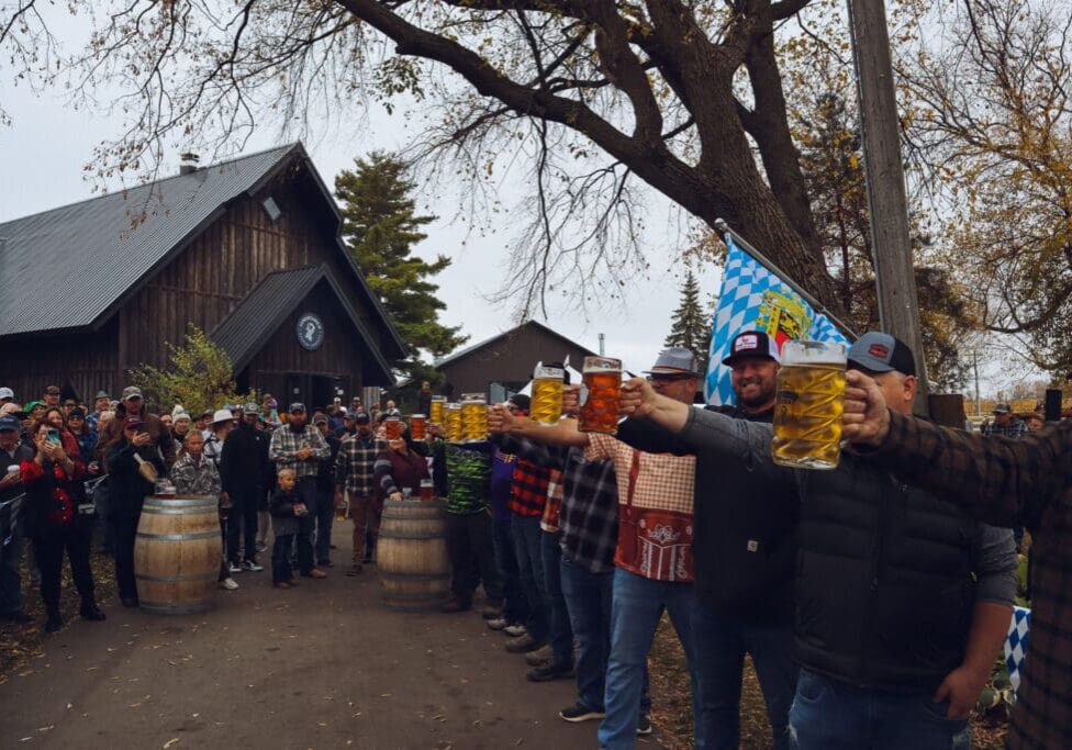 A group of people outdoors hold large beer steins in an Oktoberfest contest, cheered on by a crowd. Trees with autumn leaves and rustic buildings are in the background, capturing the festive spirit at 22 Northmen Brewing.