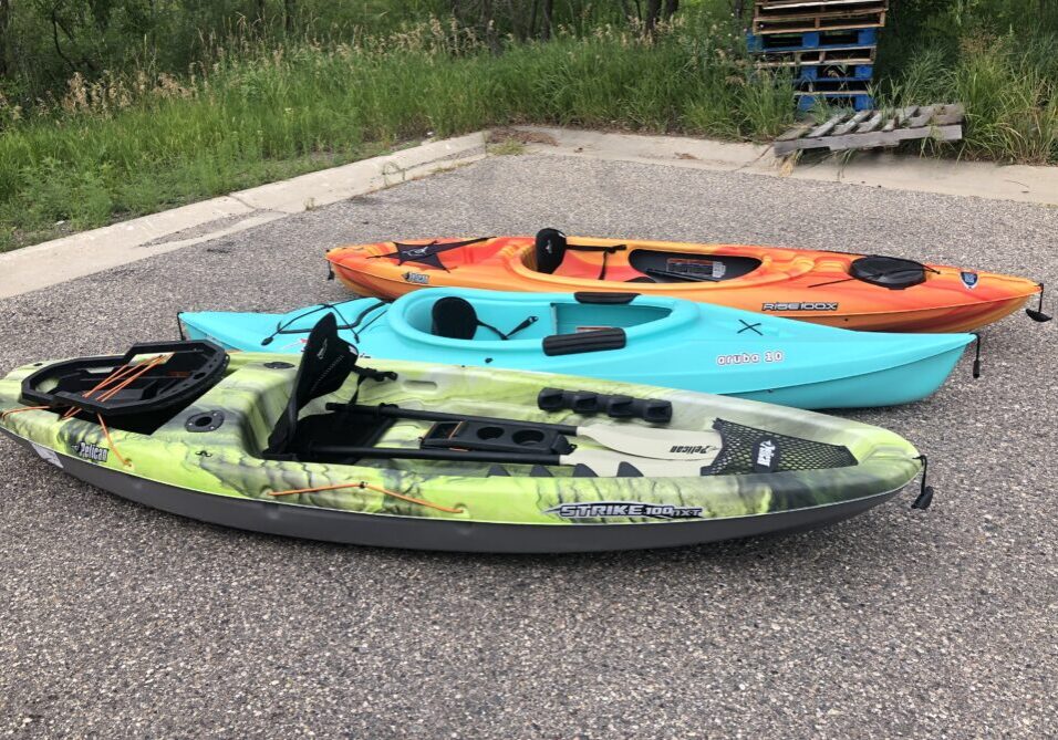 Three kayaks—one green camo, one blue, and one orange—from an Outdoor Gear Rental are parked on a gravel surface near a grassy area and wooden pallets, lined up side by side.