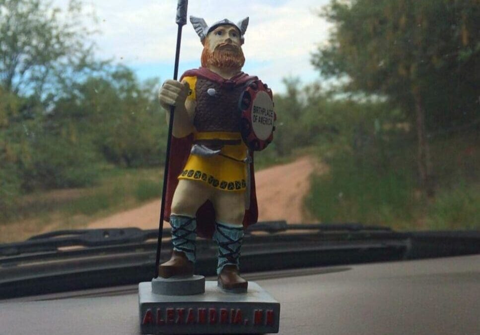 A Viking figurine with a spear and shield stands on a car dashboard, ready for the adventures of lil' ole. The base reads "ALEXANDRIA, MN." A dirt road and green trees stretch out under a cloudy sky through the windshield.