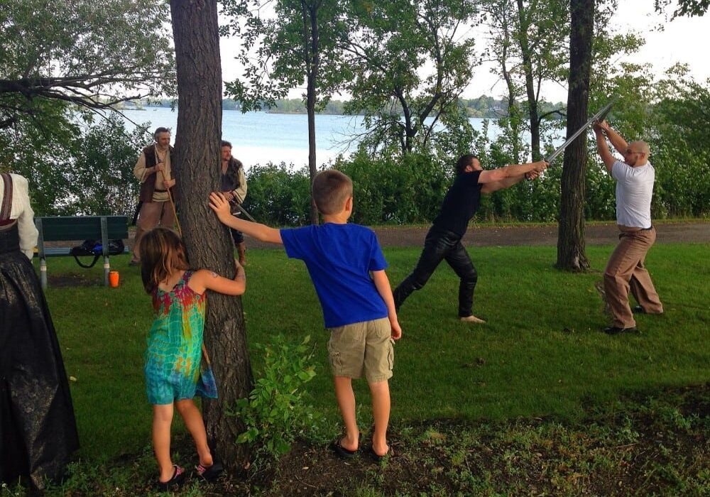 Two kids watch as two adults have a dramatic sword fight in a park near a lake, while others in costumes—suggesting a Viking adaptation or medieval fantasy event—stand nearby, reminiscent of Hamlet performed at the Alexandria Farmers Market.