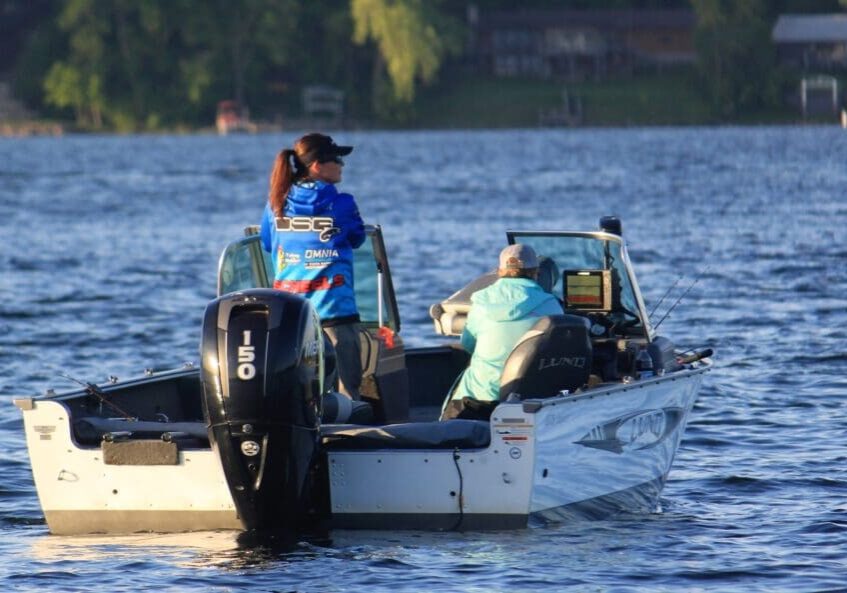 Two women anglers are fishing on a small white motorboat on a Minnesota lake. One stands near the motor while the other sits facing a screen. Trees and buildings line the distant shoreline, evoking an open water tournament atmosphere.