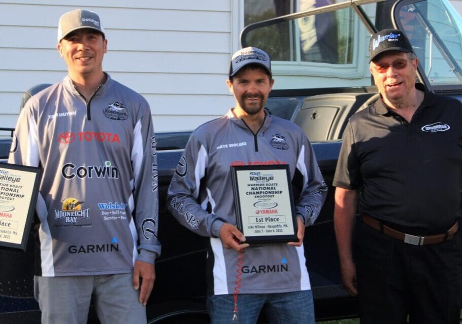Three men stand in front of a boat. Two on the left wear fishing shirts and hats, smiling and holding 1st place walleye plaques. The man on the right wears sunglasses, a cap, and a black polo shirt, also smiling.