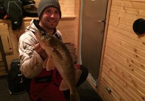 A smiling person in winter clothing holds up a large fish inside a wooden-paneled room, likely an ice fishing hut—proof of excellent fishing and great ice thickness, making for a successful day on the ice.