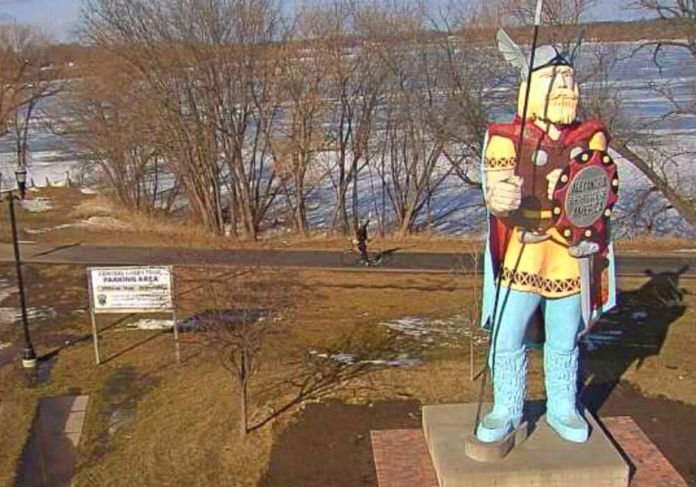A large Viking statue holding a spear and shield stands on a brick base near the walking path, offering a unique landmark for those checking central lakes trail conditions. Patchy snow covers the ground, with bare trees under a clear sky in the background.