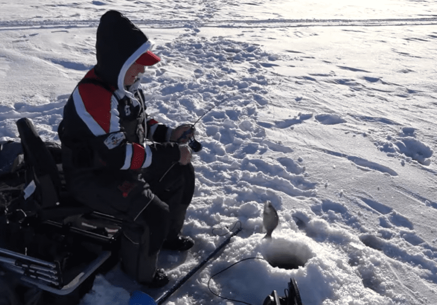 A person dressed in winter clothing sits on a chair while ice fishing, showcasing ice fishing success as they pull a fish from an ice hole on a snowy, frozen lake in the Midwest. Fishing equipment is scattered nearby.