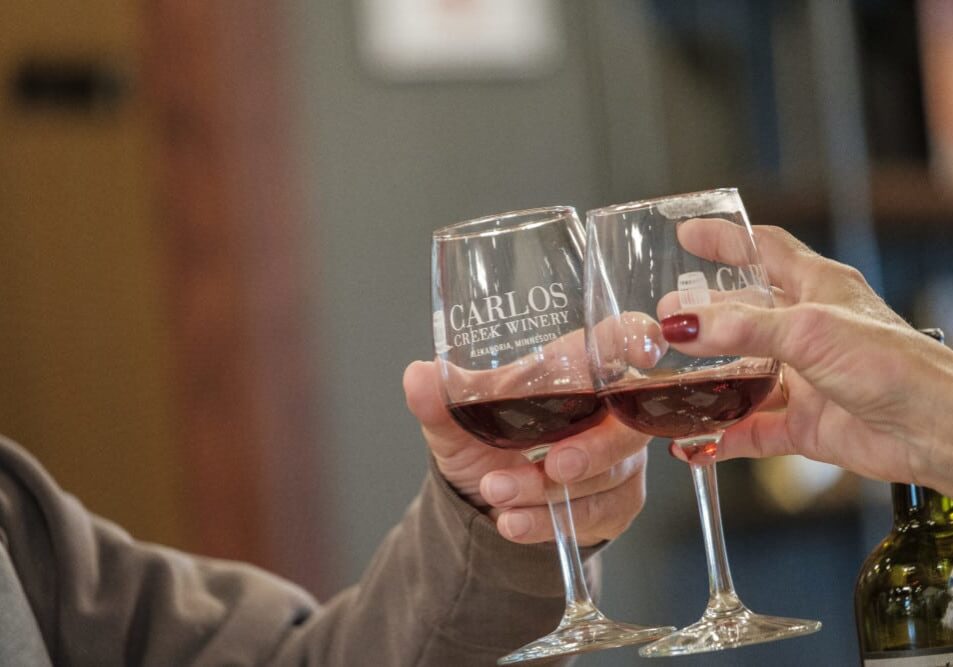 Two people clinking "Carlos Creek Winery" red wine glasses, celebrating a romantic getaway indoors—a perfect Valentine's Day moment.