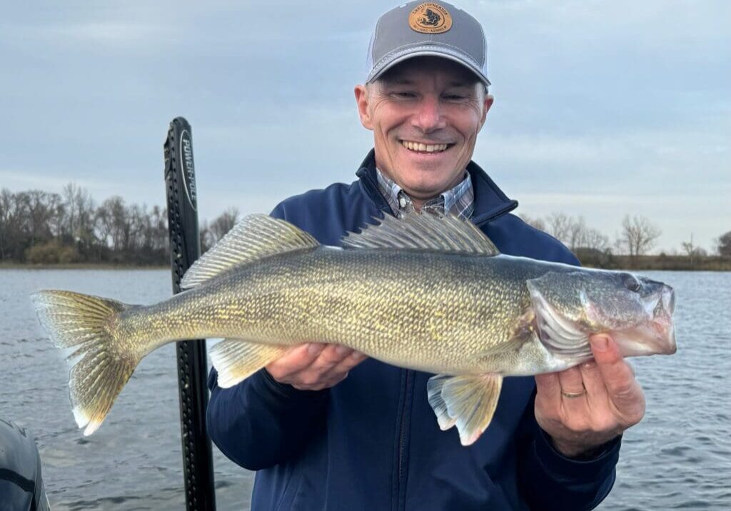 A smiling man in a blue jacket and cap holds a large fish in both hands while standing on a boat, capturing the excitement of end of the season fishing with a lake and bare trees in the background.
