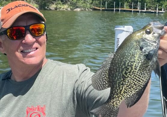 A smiling man wearing sunglasses and a cap holds up a large crappie fish on a boat, with a calm lake and shoreline in the background—a perfect day to share some crappie tips under the sun.