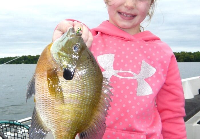 Smiling young girl in a pink hoodie holds a large fish with one hand while standing on a boat, water and trees in the background—perfect for this year’s 4th of July fishing report.