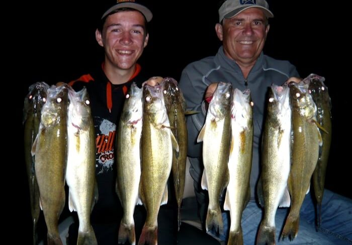 Two smiling men proudly hold up several freshly caught fish, displaying their successful summer walleye fishing haul against a dark background, likely at night. Both appear happy and content, dressed in casual outdoor clothing and caps.