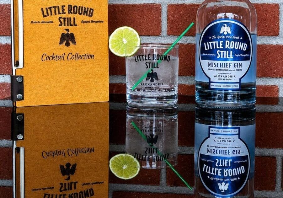 A bottle of Little Round Still Mischief Gin, a crisp gin and tonic with lime and straw, and a cocktail menu rest on a table in front of a red brick wall—setting the perfect scene for discovering the craft of Little Round Still.