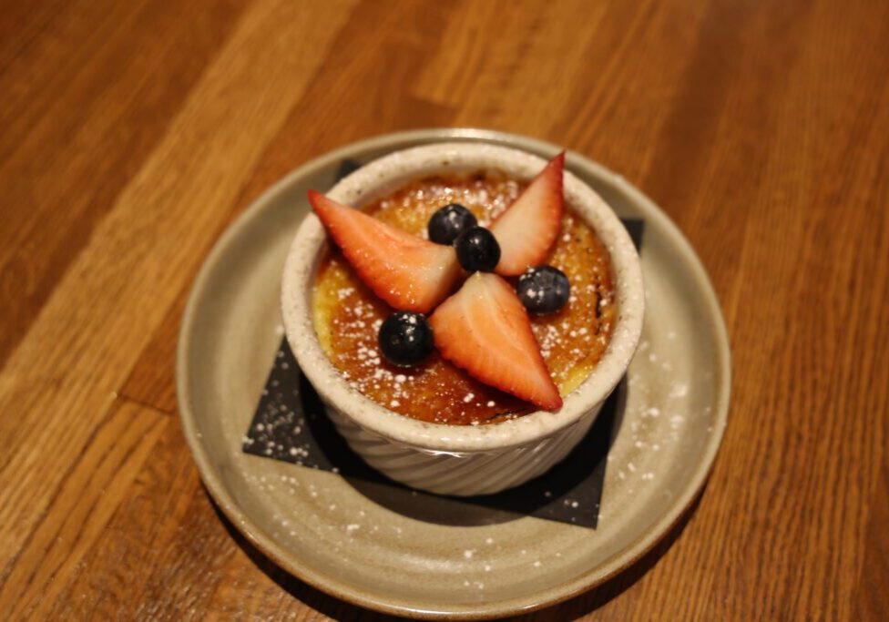 A ramekin of crème brûlée topped with strawberry slices and blueberries, lightly dusted with powdered sugar, sits on a beige plate—an elegant dessert often found at top MN Restaurants in Alexandria.