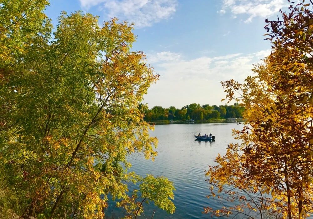 A small boat with people floats on a calm lake surrounded by trees, showing green and golden hues in this med September fall color update, beneath a partly cloudy blue sky.