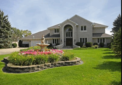 Lake Pointe Lodge is a large, two-story brick house with an arched entryway, surrounded by a well-maintained green lawn. A circular flower bed with a stone fountain sits in front, while trees gracefully frame the yard.