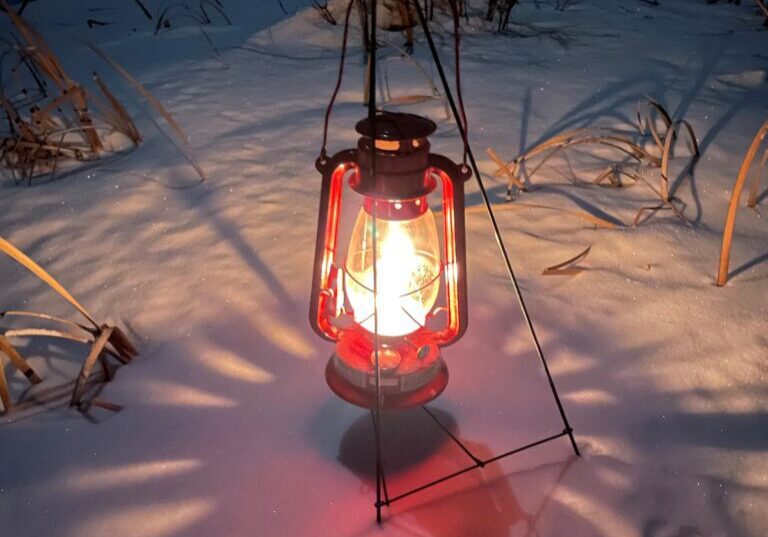 A red lantern glows warmly on snow-covered ground at dusk during a 2022 Candlelit Walk in the park, surrounded by dry grass and a dark outdoor landscape.