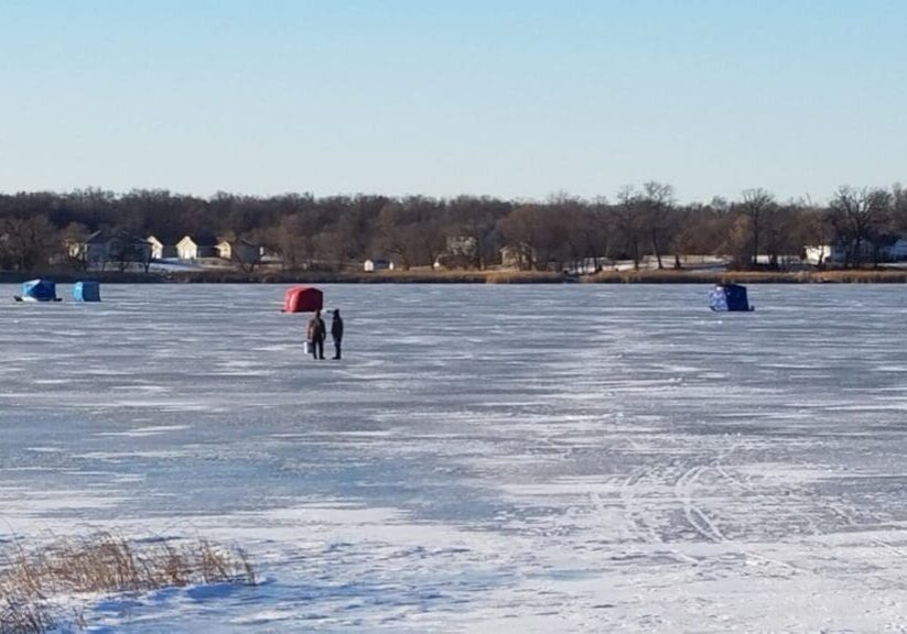 Two people walk on a frozen lake covered in snow and ice, heading toward colorful ice fishing shelters. Houses and trees line the far shore beneath a clear blue sky.