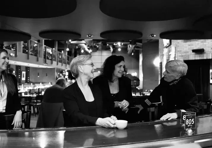 Four women sit and smile together at a curved bar counter in a modern restaurant or lounge, enjoying pleasant conversation about Songs by Boys by Girls. The scene is captured in timeless black and white.