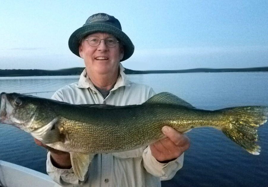 An older man wearing glasses and a bucket hat smiles while holding a freshly caught fish on a boat, with calm lake water and a distant shoreline in the background.